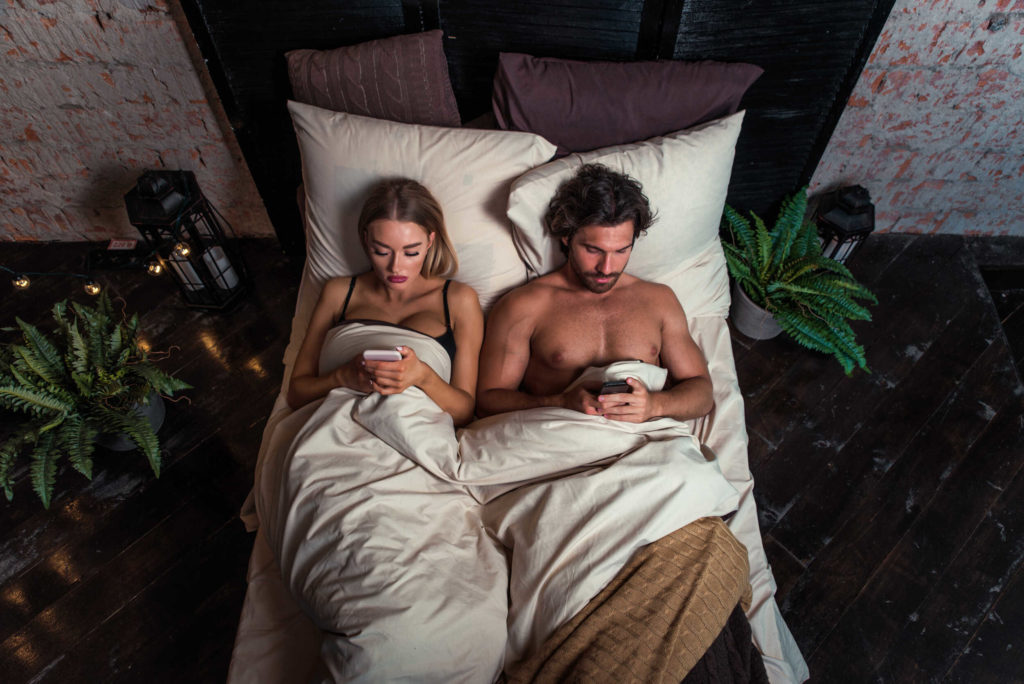 Un couple dans son lit. L'homme et la femme sont sur leurs portables respectifs. 